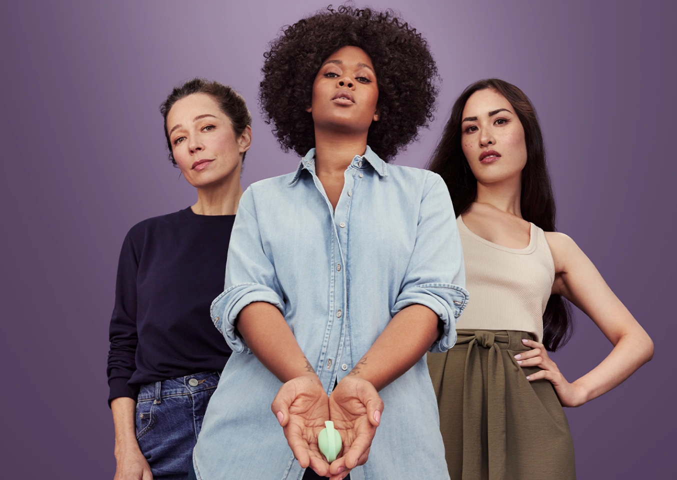 Three women standing in front of purple background. An Asian woman, in her 20s, on the left. A black woman, in her 30s, in the center, holding Elvie Trainer. A white woman, in her 50s, on the right.